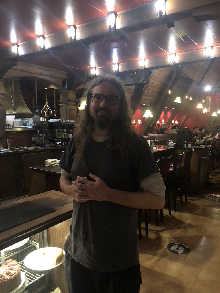 Jess Coble aka The Zero Collective stands in side a brightly lit, and architecturally strange los angeles valley deli. They are smiling, with black rim glasses a goateee and long gorgeous curly hair. 